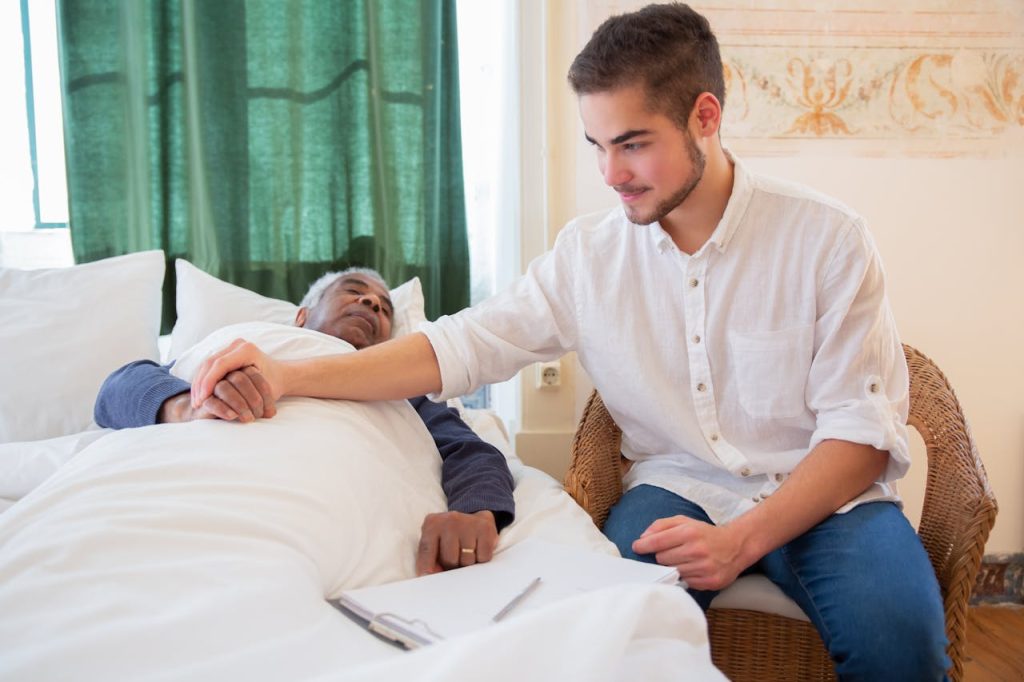 Man in White Button Up Shirt Sitting Beside Elderly Man in Bed