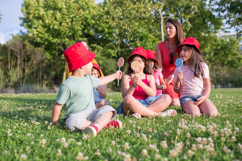 Children Sitting on Green Grass Field Holding Lollipops
